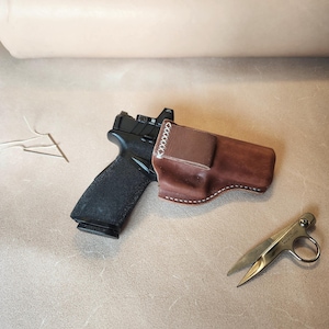 Op de afbeelding: Een zwart pistool met een bruine leren holster, witte stiksels en een kleine schaar. Het pistool is gedeeltelijk in de holster gestoken. De schaar is zilverkleurig en heeft een ring voor een vinger.