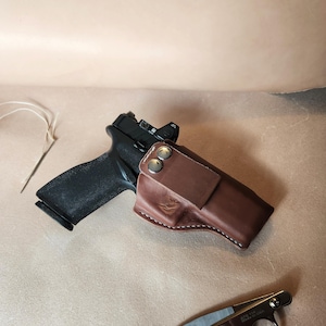 Op de afbeelding: Een zwart pistool met een rood puntvizier ligt naast een bruine leren holster. De holster is vastgezet met twee zilveren drukknopen. Een kleine schaar en een naald met draad zijn ook aanwezig.