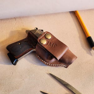 Op de afbeelding: Een bruine leren holster met witte stiksels en messing klinknagels. De holster heeft de initialen "IMP" gegraveerd op de voorkant. Een zilveren pistool zit in de holster.