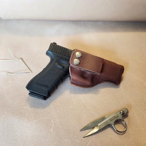 Op de afbeelding: Een zwart pistool met een bruine leren holster en een kleine zilveren schaar. De holster is vastgezet met twee zilveren klinknagels. De schaar heeft een ring voor een vinger en de tekst "ATC 114 MADE IN USA".