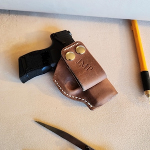 Op de afbeelding: Een bruine leren holster met witte stiksels en twee messing drukknopen. De holster is gepersonaliseerd met de initialen "JMP".