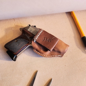 Op de afbeelding: Een bruine leren holster met witte stiksels en de initialen "JMP" in reliëf op de voorkant. Een zilveren pistool zit in de holster.
