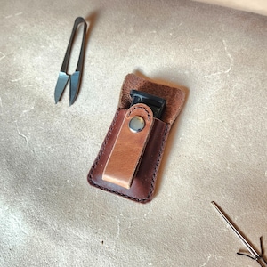 Op de afbeelding: Een handgemaakte leren etui in donkerbruin met een lichtere bruine flap, vastgezet met een zilveren drukknop. De etui bevat een zwart object. Een kleine schaar en een naald zijn ook zichtbaar.