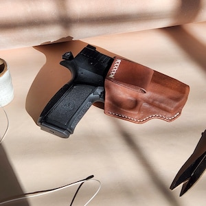 Op de afbeelding: Een zwart pistool in een bruin leren holster met witte stiksels. De holster is op maat gemaakt en lijkt nieuw te zijn. De afbeelding is gemaakt op een lichtbeige oppervlak met een spoel garen en een tang op de achtergrond.