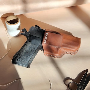 Op de afbeelding: Een zwart pistool in een bruine leren holster, met witte stiksels. Een spoel wit garen en een schaar zijn ook zichtbaar. De items zijn gerangschikt op een lichtbruin oppervlak.