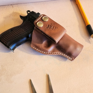 Puede incluir: Una funda de cuero marrón con las iniciales "JMP" estampadas en la parte delantera, asegurada con dos broches de color latón. Una pistola negra descansa junto a la funda. También se ven un lápiz amarillo y herramientas metálicas.