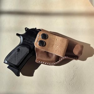 Op de afbeelding: Een zwart Bersa semi-automatisch pistool dat gedeeltelijk in een bruine lederen holster zit. De holster is voorzien van een lichtere beige lederen riemclip die is vastgezet met twee zilverkleurige metalen drukknopen en witte stiksels.