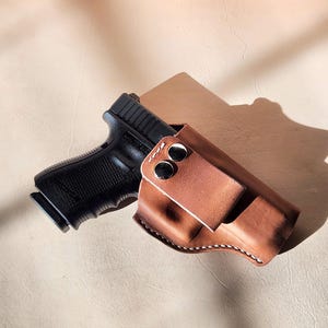 Op de afbeelding: Een zwart pistool naast een bruine leren holster. De holster heeft een riemclip met twee zilveren drukknopen en witte stiksels langs de randen. De achtergrond is lichtbruin.