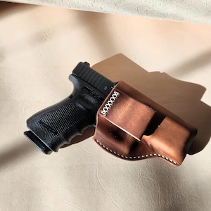 Op de afbeelding: Een zwart pistool gedeeltelijk in een bruine leren holster gestoken. De holster is gestikt met witte draad en heeft een gebogen vorm. De achtergrond is lichtbruin.