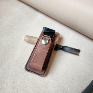 Może przedstawiać: Brązowy skórzany uchwyt na narzędzia z zapięciem na zatrzask, trzymający czarne i srebrne narzędzie do rzeźbienia. Uchwyt jest zszyty ciemną nicią i leży na jasnobrązowej powierzchni. Widoczne jest również narzędzie z drewnianym uchwytem.