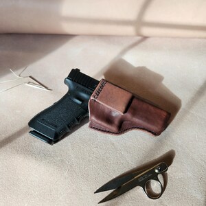 Op de afbeelding: Een zwart pistool naast een bruine leren holster en een kleine schaar. De holster is gestikt met een contrasterende draad. De schaar is zilverkleurig met een ring voor een vinger. Een naald en draad zijn ook zichtbaar.
