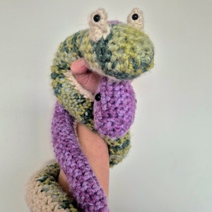 Puede incluir: Un juguete de serpiente de crochet en color verde, morado y blanco. La serpiente tiene ojos grandes y negros y una expresión amigable.