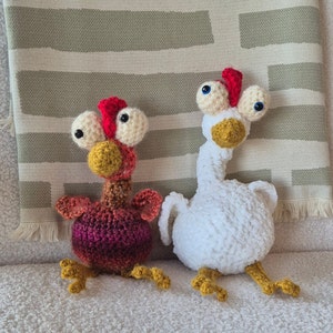 May include: Two crocheted chicken toys. One is white with large blue eyes and a yellow beak. The other is red, brown, and yellow with smaller blue eyes and a yellow beak.