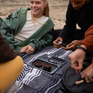 May include: A group of friends are gathered around a black rolling tray with two metal grinders. The tray has a built-in poker and a small compartment for storing accessories. The friends are sitting on a blanket with a black and white pattern.