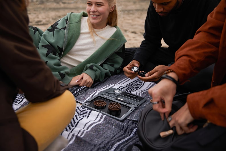 May include: A group of friends are gathered around a black rolling tray with two metal grinders. The tray has a built-in poker and a small compartment for storing accessories. The friends are sitting on a blanket with a black and white pattern.