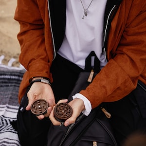 May include: Two wooden grinder pieces, one open and one closed, are held in the hands of a person wearing a brown jacket and a white shirt. The person is sitting on a black and white blanket. A black backpack with a brown zipper pull is in the background.