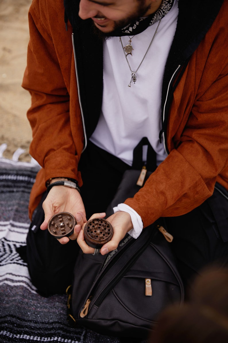 May include: Two wooden grinder pieces, one open and one closed, are held in the hands of a person wearing a brown jacket and a white shirt. The person is sitting on a black and white blanket. A black backpack with a brown zipper pull is in the background.