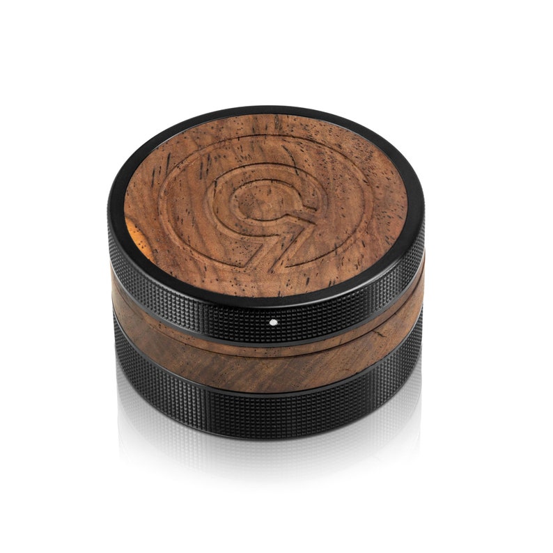 May include: A round wooden and black speaker with a textured surface and a small white dot on the front. The speaker has a wood grain pattern and a black metal ring around the top and bottom.
