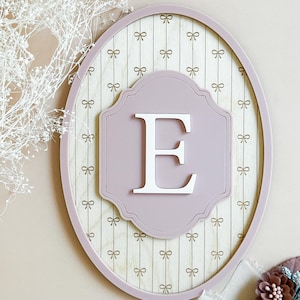 May include: A decorative wooden wall hanging with a pale pink oval frame. The interior has a light wood grain pattern with small bow accents. A pale pink plaque with the letter "E" is centered.