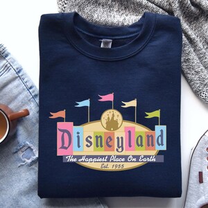 A navy blue sweatshirt with a Disneyland logo, a pair of jeans, a gray sweater, a cup of coffee, and a pair of sneakers are arranged on a white surface.