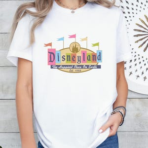 A woman wearing a white t-shirt with a colorful graphic of Disneyland and the text "Disneyland The Happiest Place On Earth" printed on it.