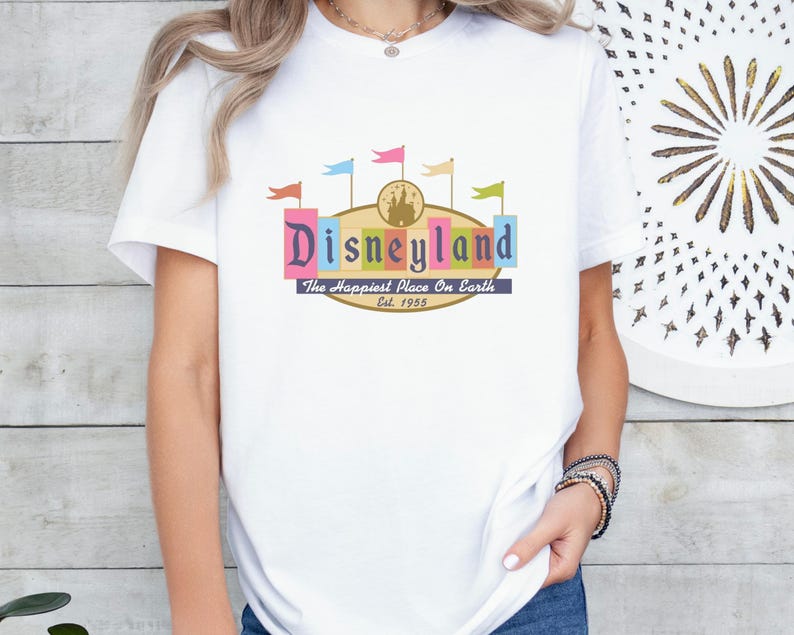 A woman wearing a white t-shirt with a colorful graphic of Disneyland and the text "Disneyland The Happiest Place On Earth" printed on it.