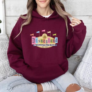 A woman wearing a burgundy hoodie with a Disneyland logo is sitting on a couch.
