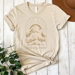 May include: Beige t-shirt with a graphic of clouds and sun rays inside an arch. The text "STORMBLESSED" is below the graphic. The shirt is folded and knotted at the bottom. The background is a light wood surface.