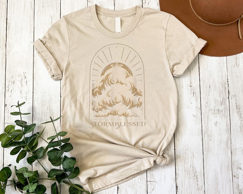 May include: Beige t-shirt with a graphic of clouds and sun rays inside an arch. The text "STORMBLESSED" is below the graphic. The shirt is folded and knotted at the bottom. The background is a light wood surface.