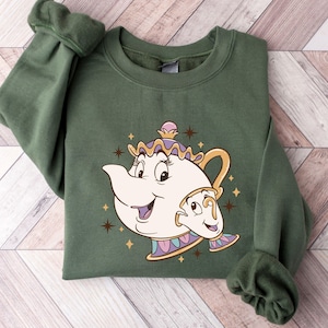 May include: An olive green sweatshirt featuring a cartoon illustration of Mrs. Potts and Chip from Beauty and the Beast. The design includes a smiling teapot and teacup with gold accents and star details. The sweatshirt is folded on a wooden surface.