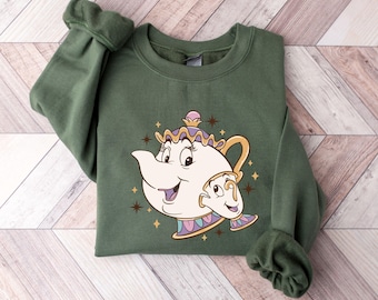 May include: An olive green sweatshirt featuring a cartoon illustration of Mrs. Potts and Chip from Beauty and the Beast. The design includes a smiling teapot and teacup with gold accents and star details. The sweatshirt is folded on a wooden surface.