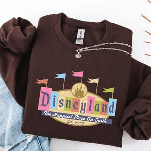A brown sweatshirt with a Disneyland logo and text, along with a denim jacket and some leaves, is displayed on a white background.