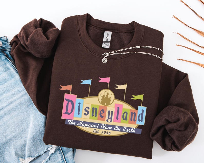 A brown sweatshirt with a Disneyland logo and text, along with a denim jacket and some leaves, is displayed on a white background.