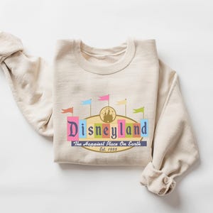 A beige sweater with a colorful graphic design featuring the text "Disneyland" and the phrase "The Happiest Place on Earth" printed on it.