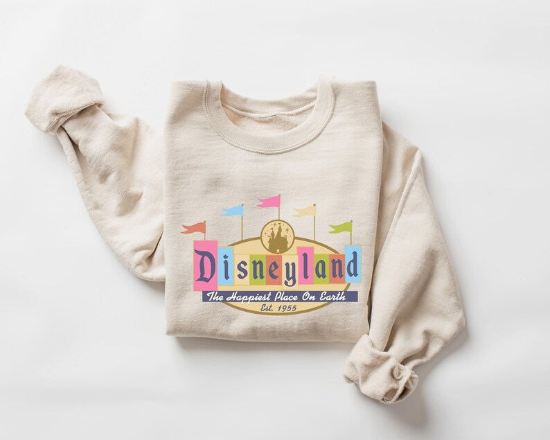 A beige sweater with a colorful graphic design featuring the text "Disneyland" and the phrase "The Happiest Place on Earth" printed on it.