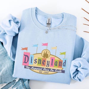 A light blue t-shirt with a Disneyland logo and text, along with a denim jacket and some leaves, is displayed on a white background.