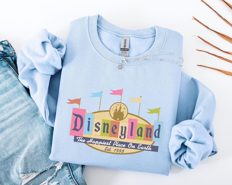A light blue t-shirt with a Disneyland logo and text, along with a denim jacket and some leaves, is displayed on a white background.