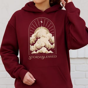 May include: Burgundy hoodie with a front pocket and a drawstring hood. The graphic on the front features a stylized illustration of clouds, a figure, and the text "STORMBLESSED" in a serif font.