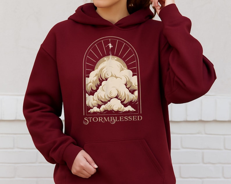 May include: Burgundy hoodie with a front pocket and a drawstring hood. The graphic on the front features a stylized illustration of clouds, a figure, and the text "STORMBLESSED" in a serif font.