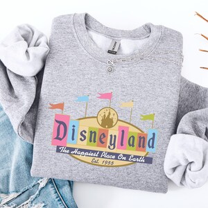 A gray sweatshirt with a Disneyland logo and text, along with a denim jacket and some orange and brown twigs, are displayed on a white background.