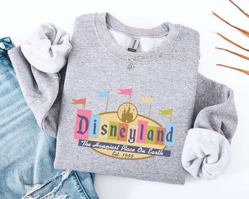 A gray sweatshirt with a Disneyland logo and text, along with a denim jacket and some orange and brown twigs, are displayed on a white background.