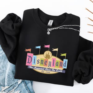 A black sweatshirt with a colorful graphic of Disneyland printed on it, along with a denim skirt and a necklace.