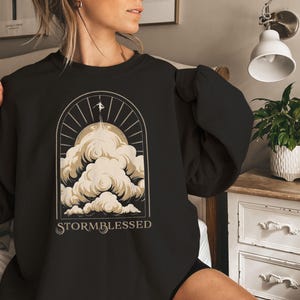 May include: Black sweatshirt with a graphic design featuring a person ascending through clouds towards a sunburst. The word "STORMBLESSED" is printed below the design in a stylized font. The sweatshirt is made of a soft material.