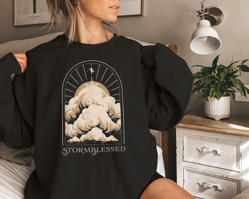 May include: Black sweatshirt with a graphic design featuring a person ascending through clouds towards a sunburst. The word "STORMBLESSED" is printed below the design in a stylized font. The sweatshirt is made of a soft material.