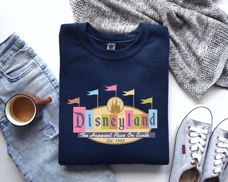 A navy blue sweatshirt with a Disneyland logo, a pair of jeans, a gray sweater, a cup of coffee, and a pair of sneakers are arranged on a white surface.