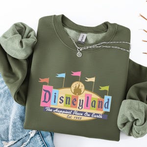 A green sweatshirt with a Disneyland logo and text, along with a denim jacket and a necklace, is displayed on a white background.