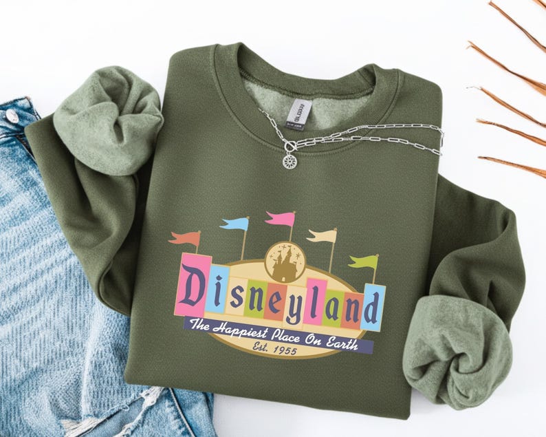 A green sweatshirt with a Disneyland logo and text, along with a denim jacket and a necklace, is displayed on a white background.