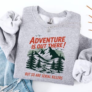 May include: A gray sweatshirt with the text "ADVENTURE IS OUT THERE! BUT SO ARE SERIAL KILLERS" in red and black. The graphic includes mountains, trees, and birds. The sweatshirt is folded on top of blue jeans.
