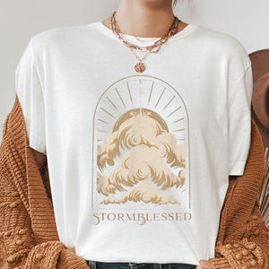 May include: White t-shirt with a gold-colored graphic of clouds and sun rays inside an arch. The text "STORMBLESSED" is below the graphic. The shirt is part of a clothing collection.