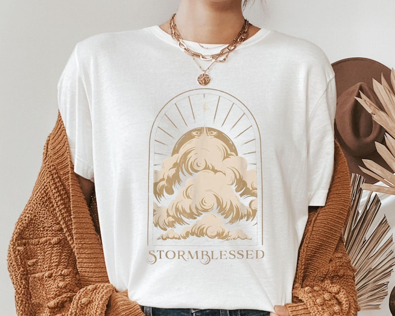 May include: White t-shirt with a gold-colored graphic of clouds and sun rays inside an arch. The text "STORMBLESSED" is below the graphic. The shirt is part of a clothing collection.